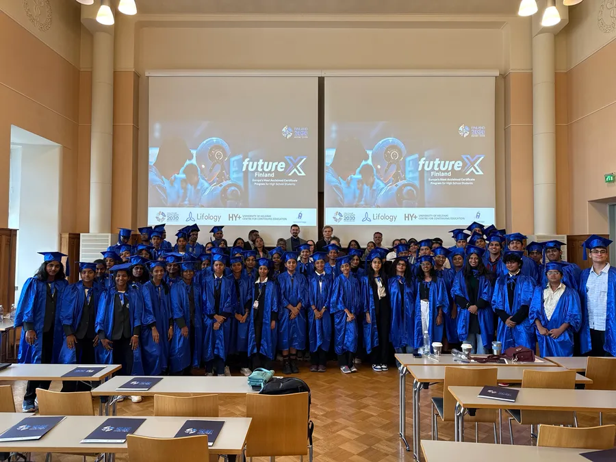 A large student group in blue graduation gowns and caps in a hall, in front of two screens showing the program title and partner logos.