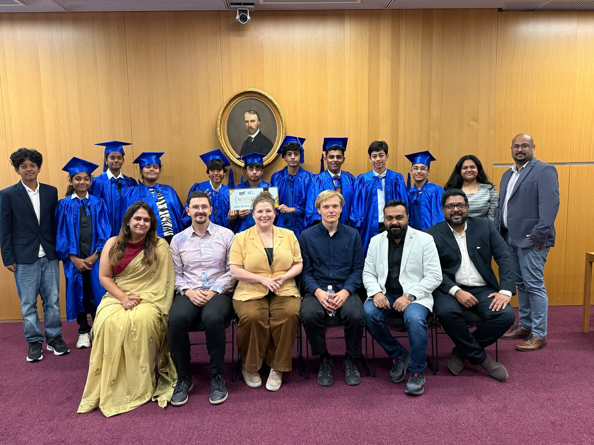Students in blue graduation gowns and caps stand behind a seated jury, one student holding a winner's certificate.