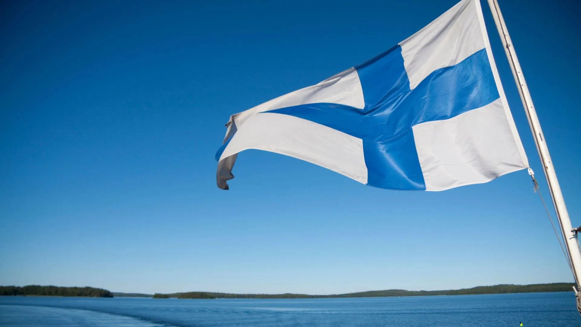 The Finnish flag flying over the sea, with forested islands on the horizon.