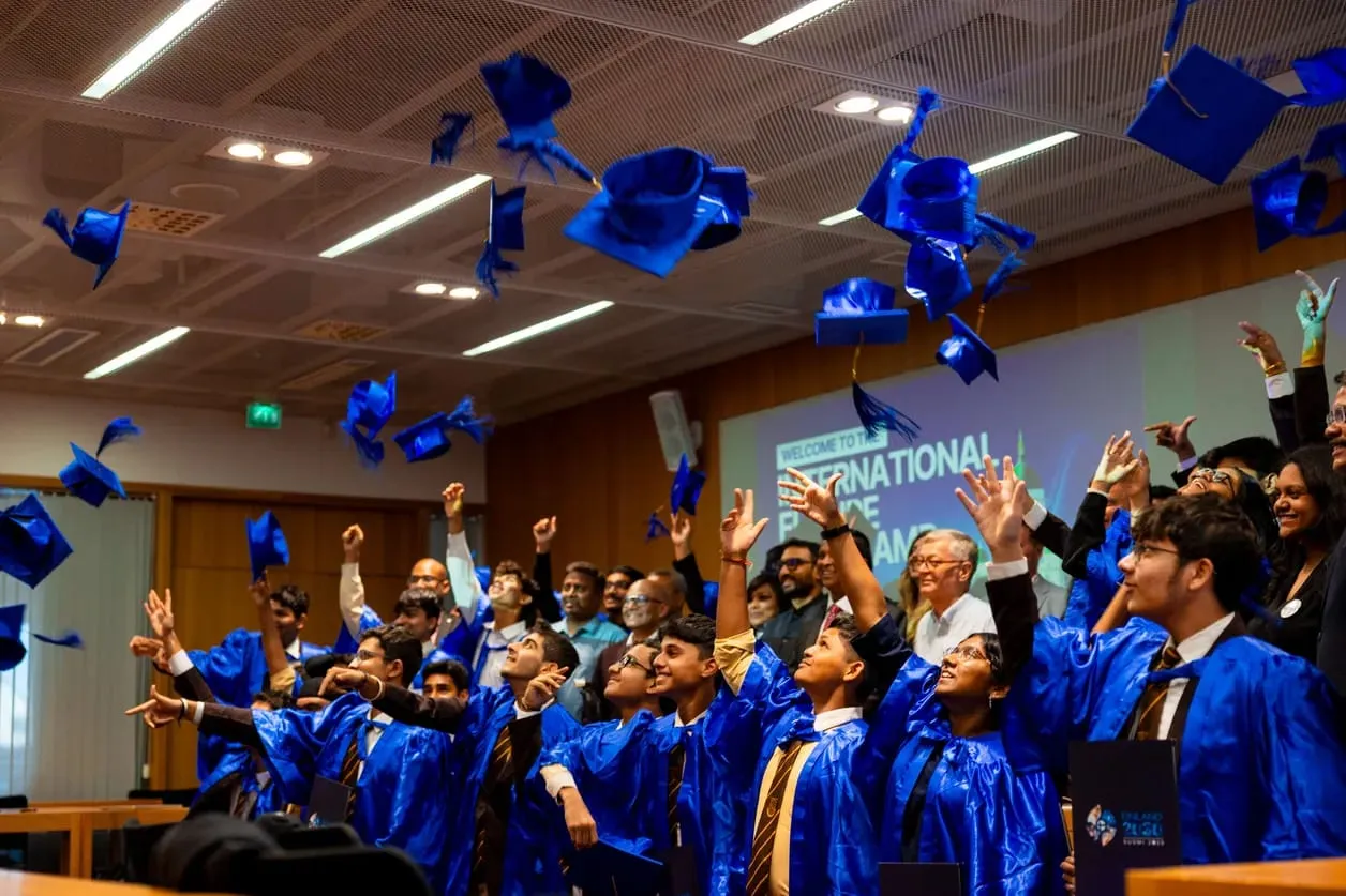 Students in blue graduation gowns throw their caps in the air in a lecture hall at the end of a program in Finland.
