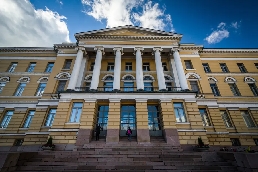 Helsingin yliopiston pylväikköinen päärakennus sinistä taivasta vasten portaiden juurelta kuvattuna.